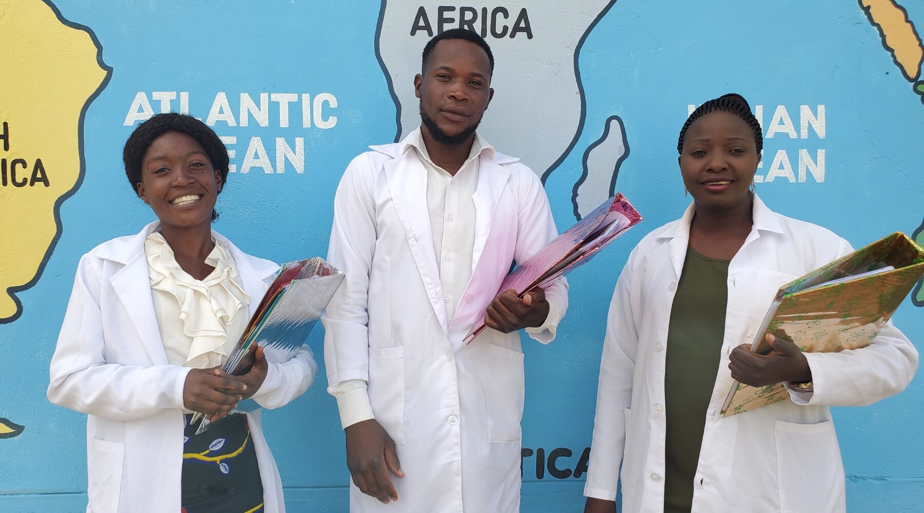 Three young adults wearing lab coats, holding binders and smiling at the camera