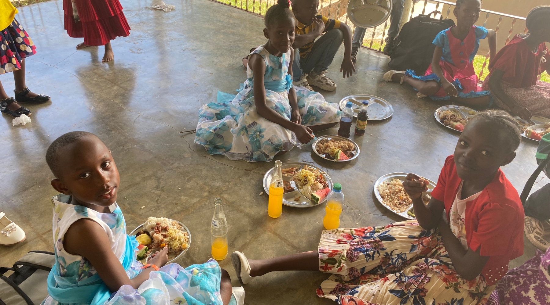 Children sitting on the ground eating a meal and drinking orange soda. 