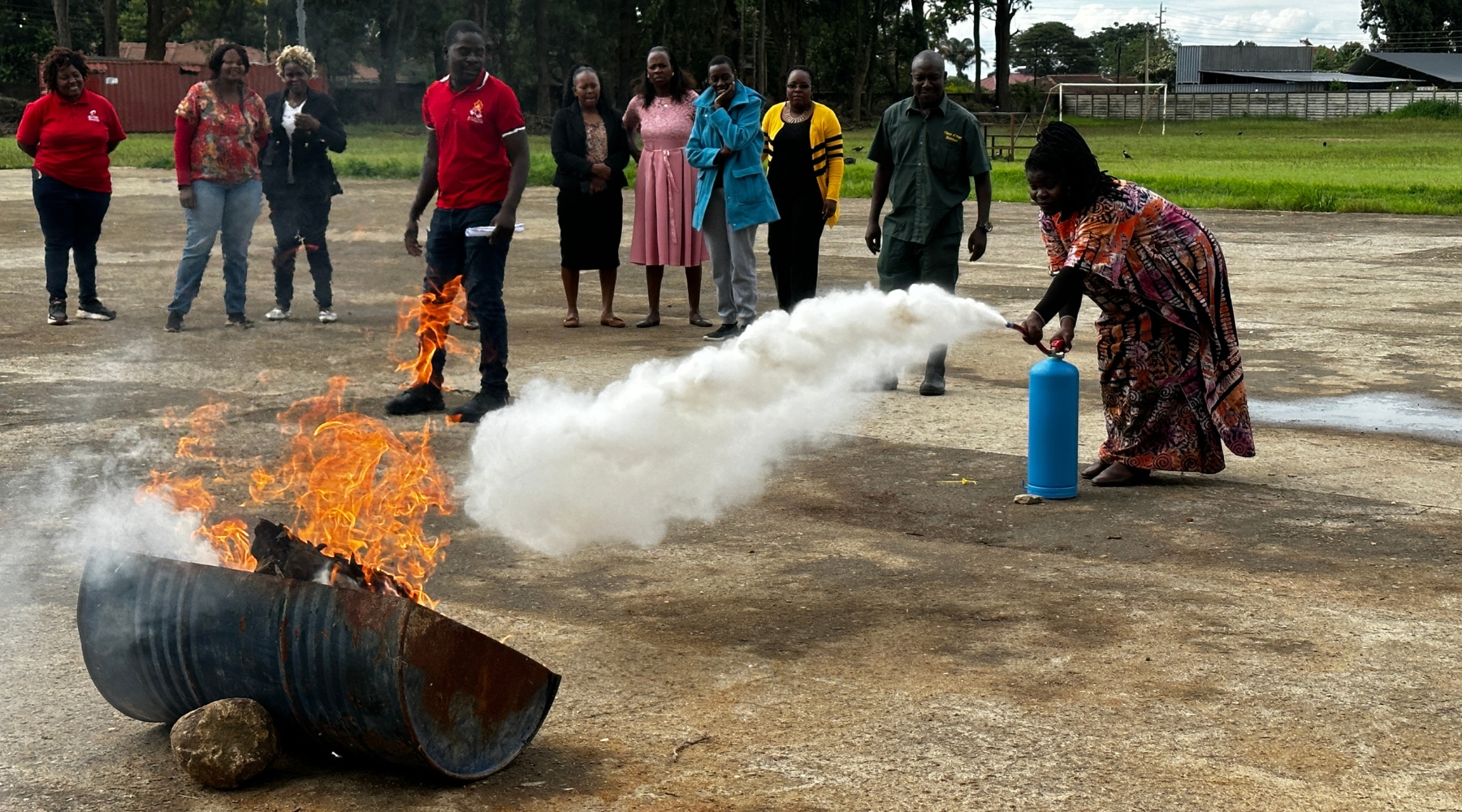 An adult holding a fire extinguisher and putting out a fire on a barrel with others watching. 