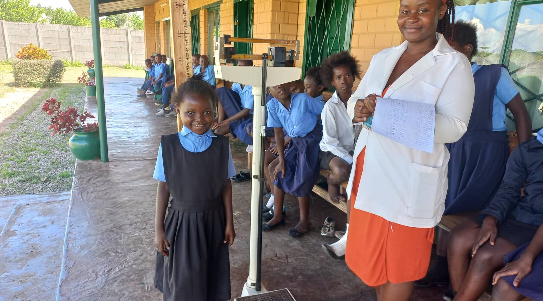 A smiling child being measured by an adult with a group of children in a line watching.