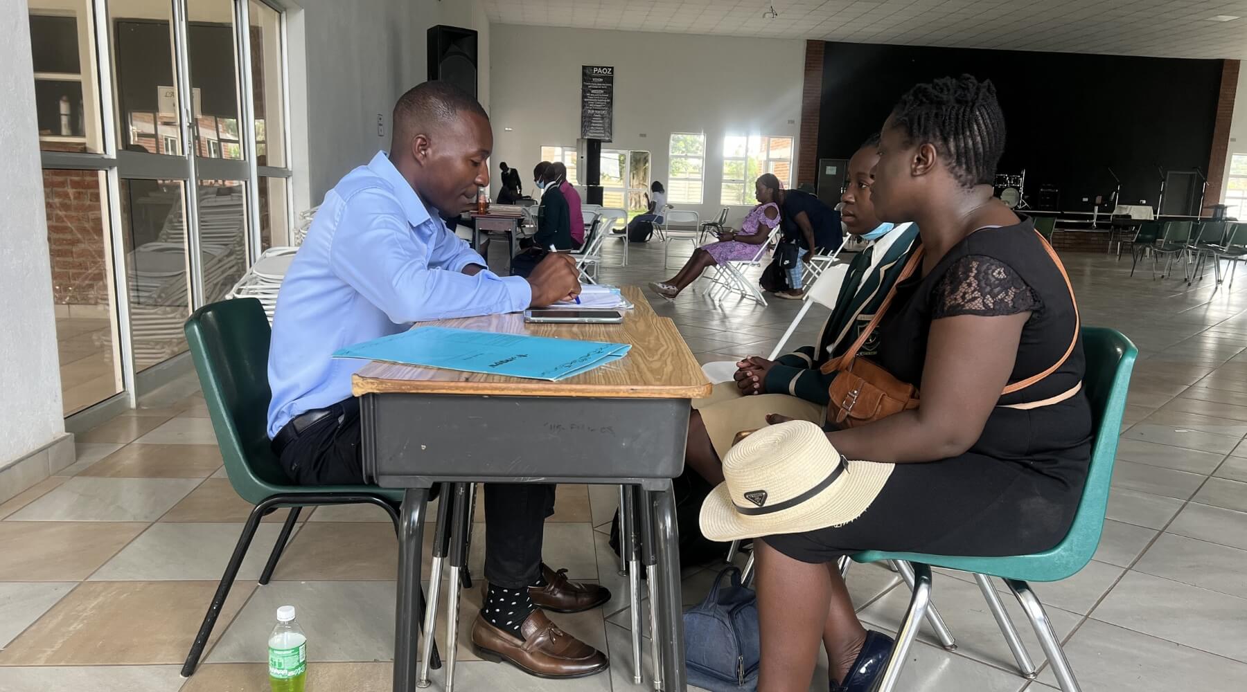An adult with a child sitting at a desk talking with a school staff member.