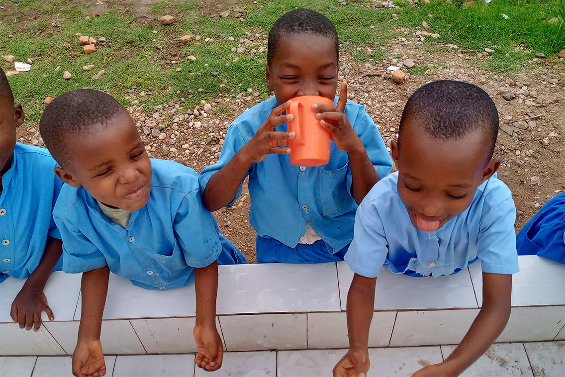 Children standing at an outdoor sink playing with and drinking water while smiling.