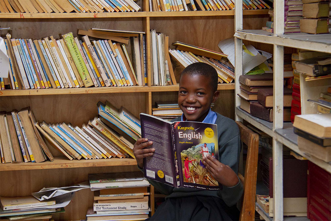A child sitting in a library, surrounded by bookshelves, holding a book and smiling at the camera.