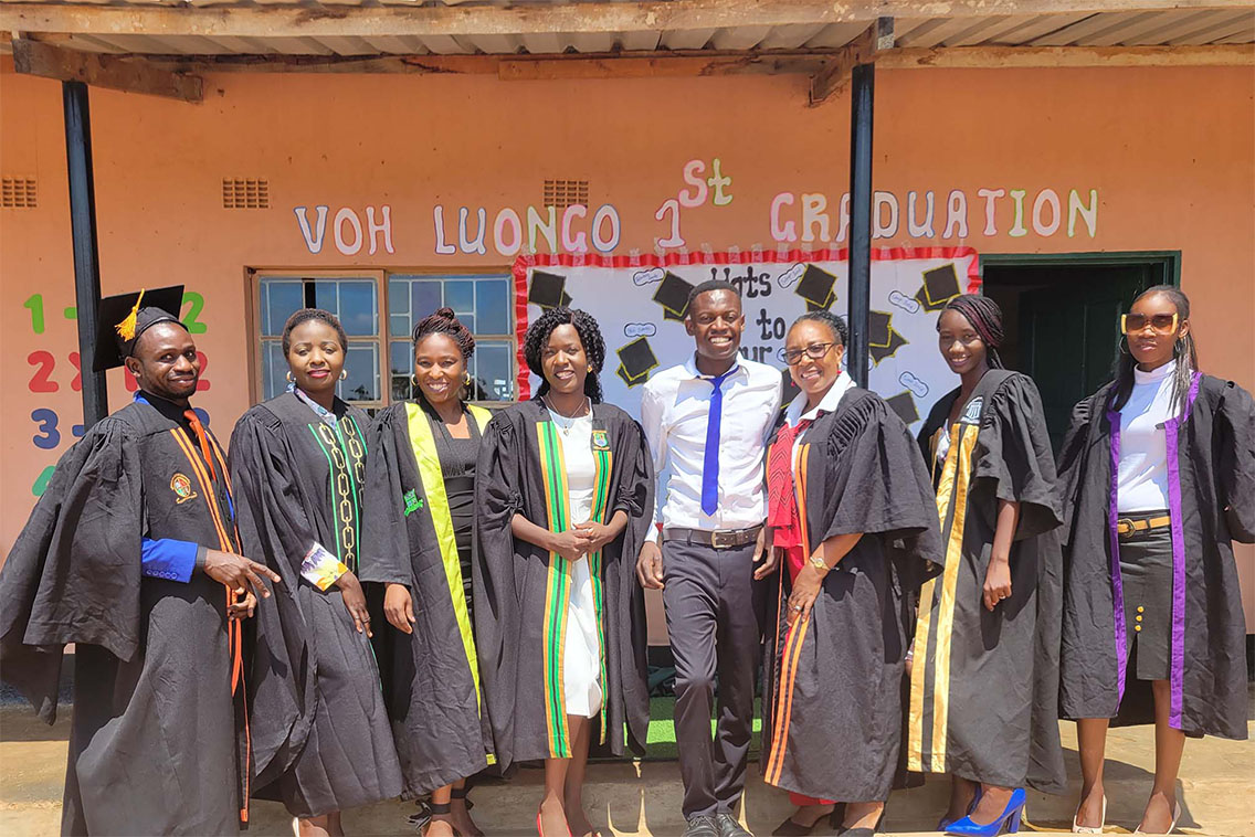 A group of young people wearing graduation gowns with "VOH Luongo 1st Graduation" sign in the background.